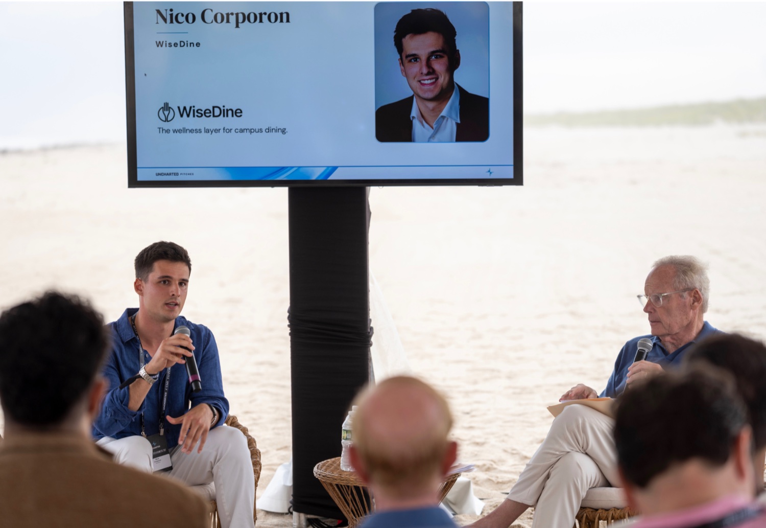 Nico Corporon, founder of WiseDine, speaking with a microphone at a pitch event; the screen behind him reads &ldquo;Nico Corporon &mdash; WiseDine&rdquo; above the WiseDine logo and the line &ldquo;The wellness layer for campus dining.&rdquo;
